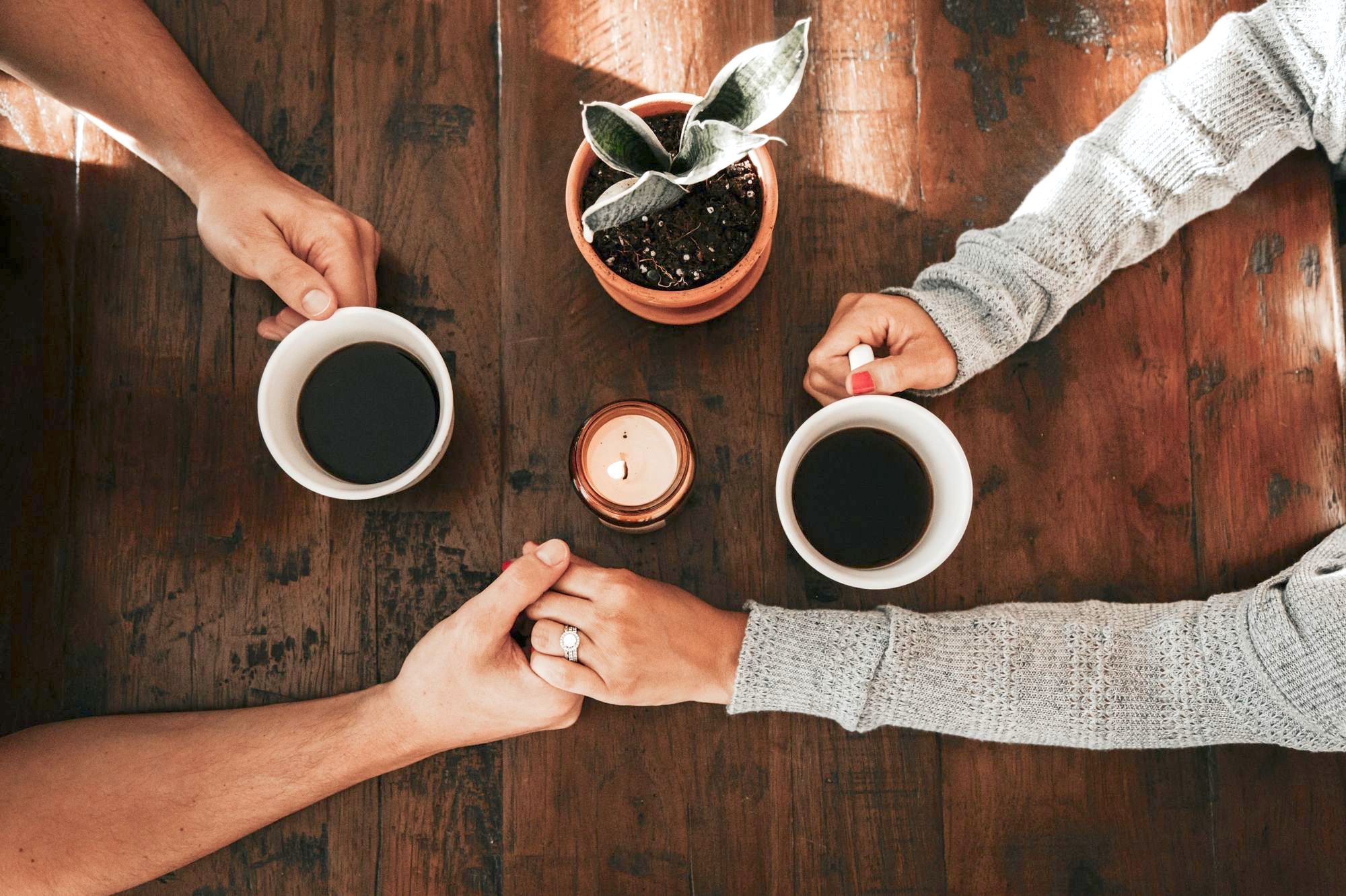 The hands of a medium skinned couple hold each other across a table. One of the hands has an engagement ring.