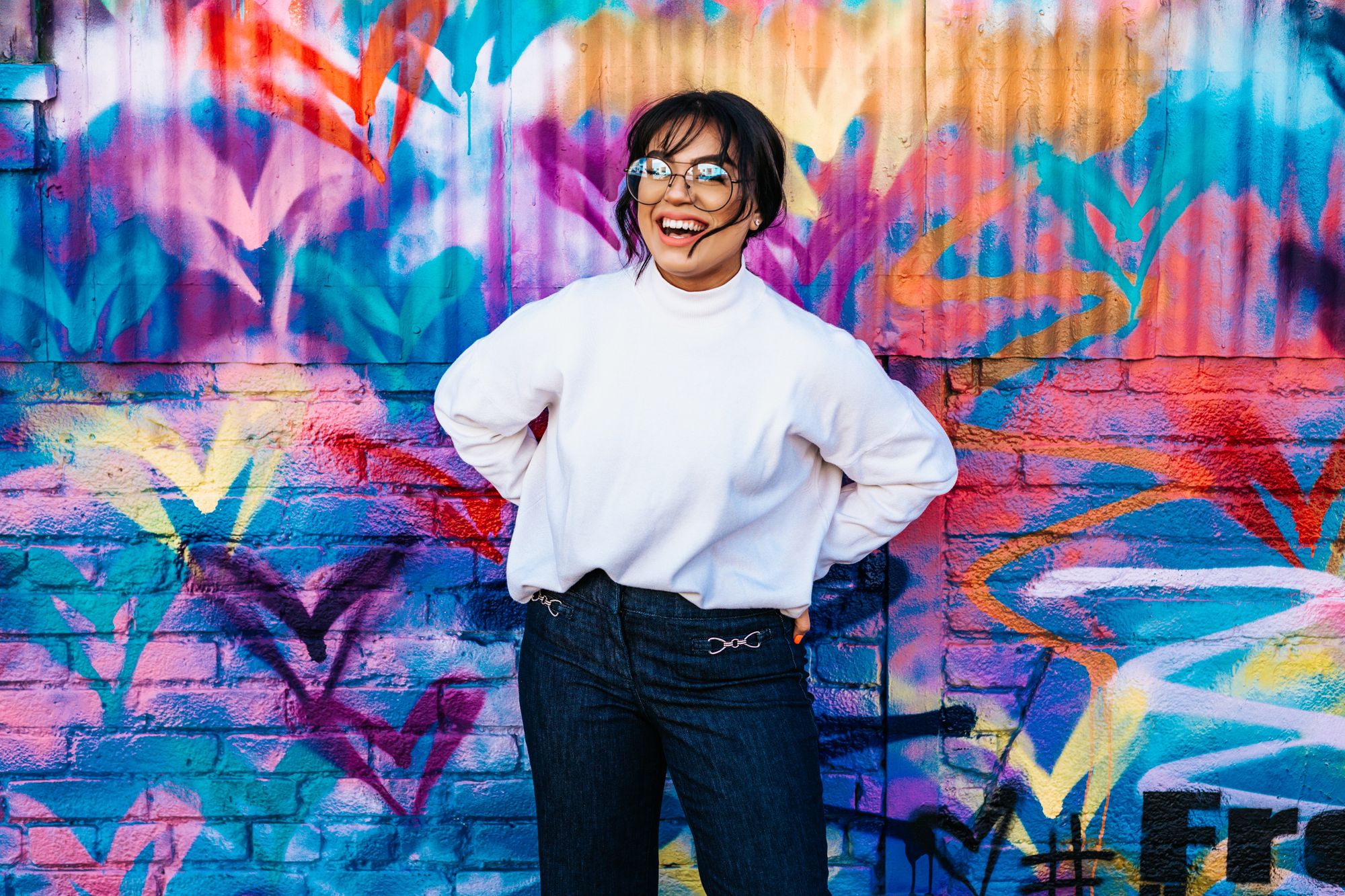 A medium skinned adult woman standing in front of a wall graffitied with hearts 