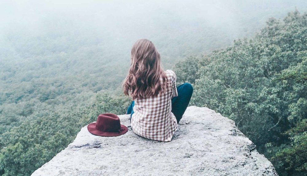 A adult with long hair, sitting on a rock, looking at a forest valley