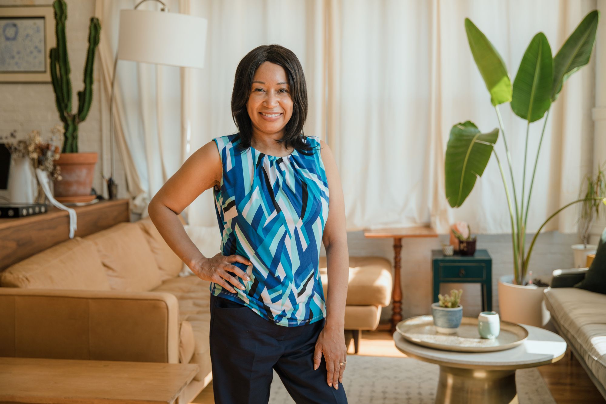 A dark skinned adult with medium length hair standing in a living room with one hand on their hip