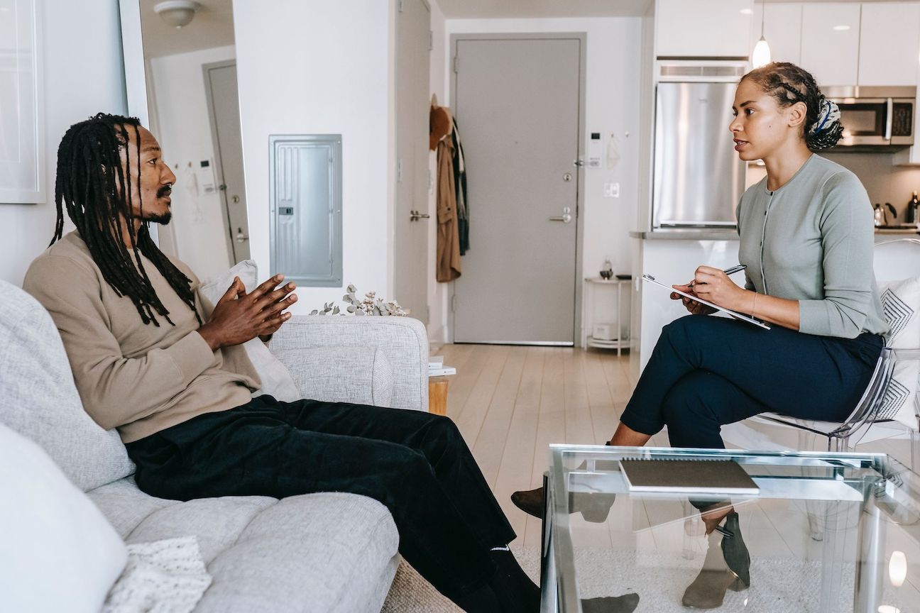 A dark skinned adult sits on a couch across from another dark skinned adult sitting in a chair taking notes