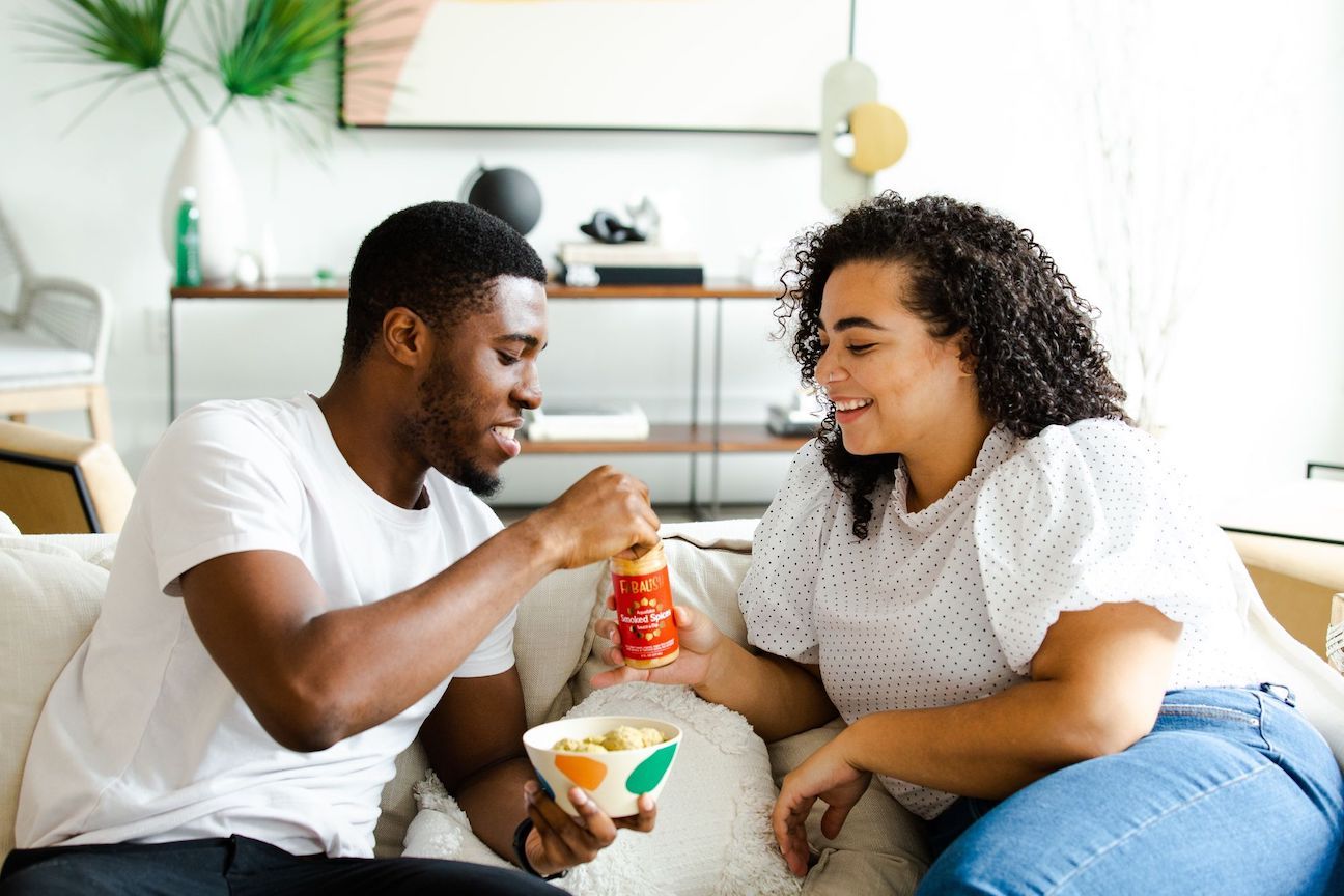 A dark skinned adult male and a medium skinned adult woman sitting on a couch together, eating