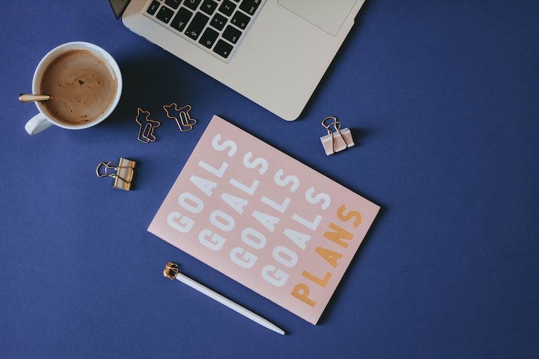 Notebook that says goals and plans on it, on a table next to a cup of coffee and a laptop