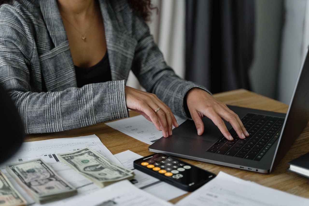 Medium skinned woman sitting at a table with a laptop, documents, cash and a calculator
