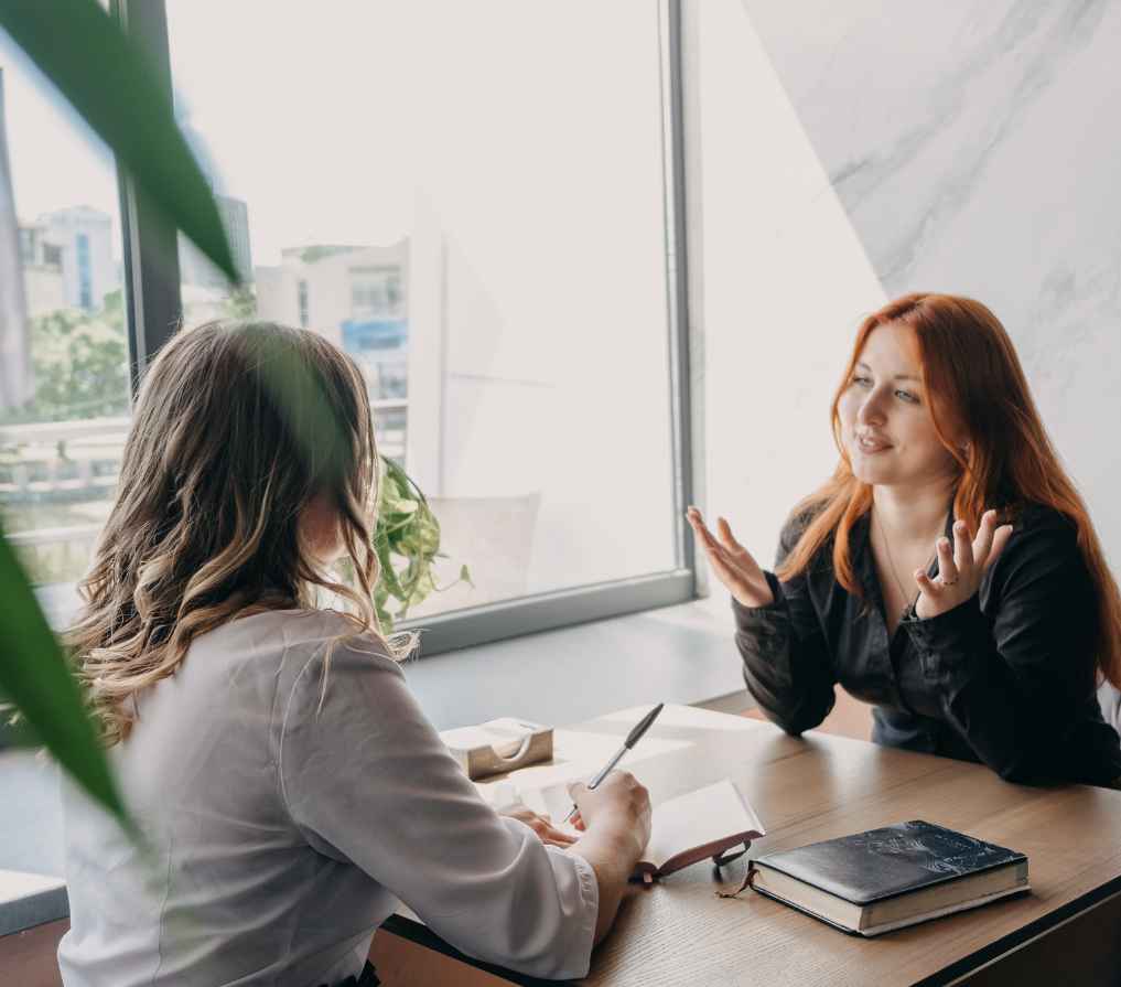 Caucasian therapist listening while a Caucasian patient speaks. Both are sitting at a table by a window.