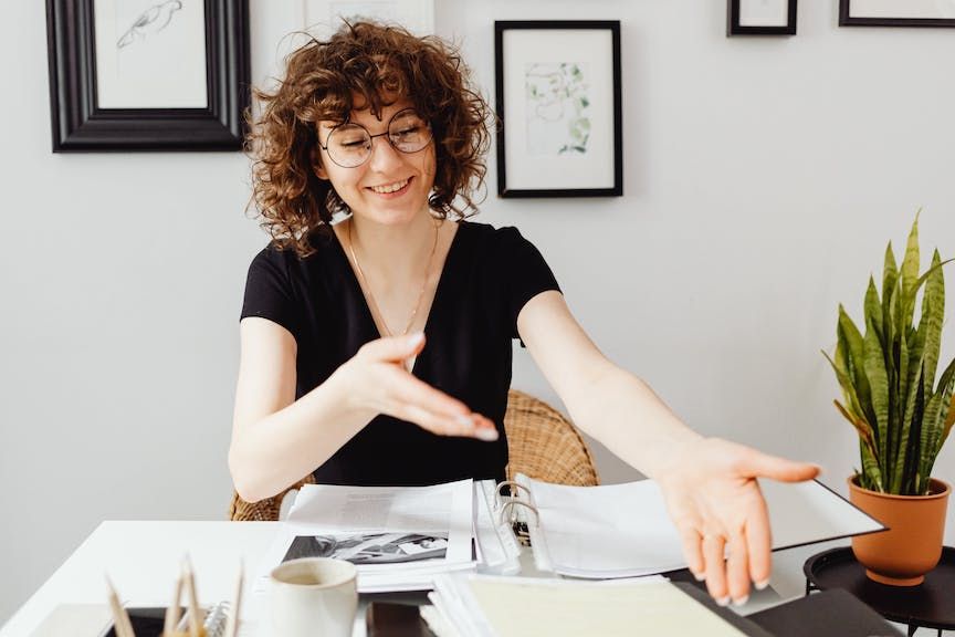 Person with curly hair and glasses sitting at a desk with binder and other paperwork pointing to it.