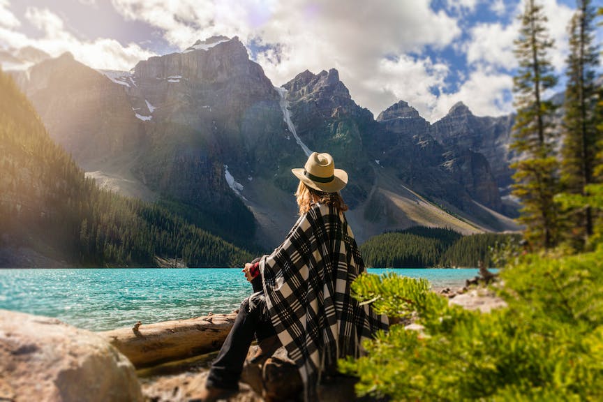 Person sitting on a log looking out at a beautiful nature scape with a lake, mountains, and the sunshining
