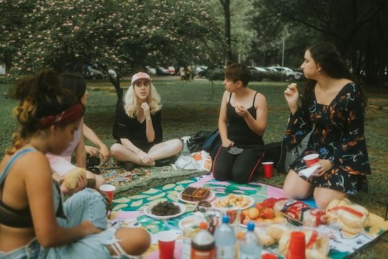 a group of adult friends sitting at a picnic together, eating food and talking