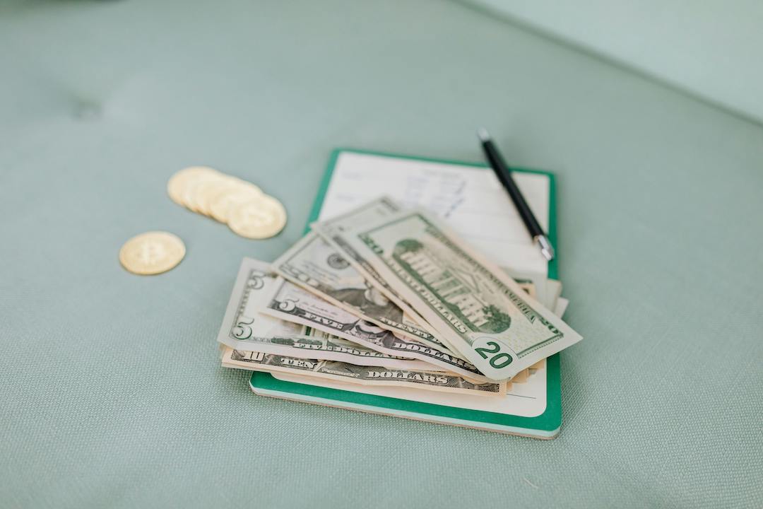 Cash and coins on top of a small notebook and pen, laying on a sage green couch.