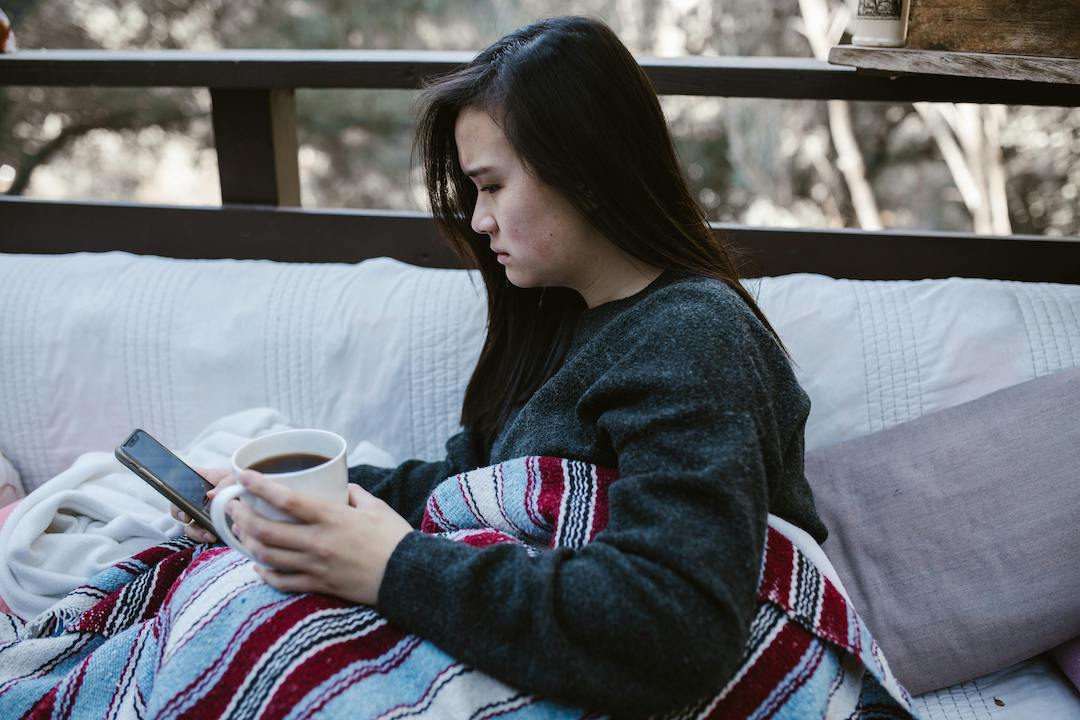 Person with light skin and black hair sitting on a cushion outside, wrapped in a blanket, holding coffee and looking at their phone with sadness in their expression.