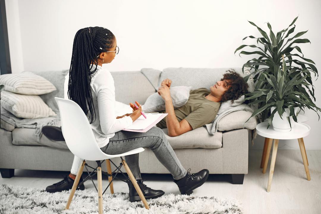 African American person with long black braids, white top, grey denim, and black boots sitting in a chair with clipboard and pen in lap. This person is looking towards a Caucasian person wearing a green tee and white denim, who is laying on a coach with a pillow on their chest.             .n