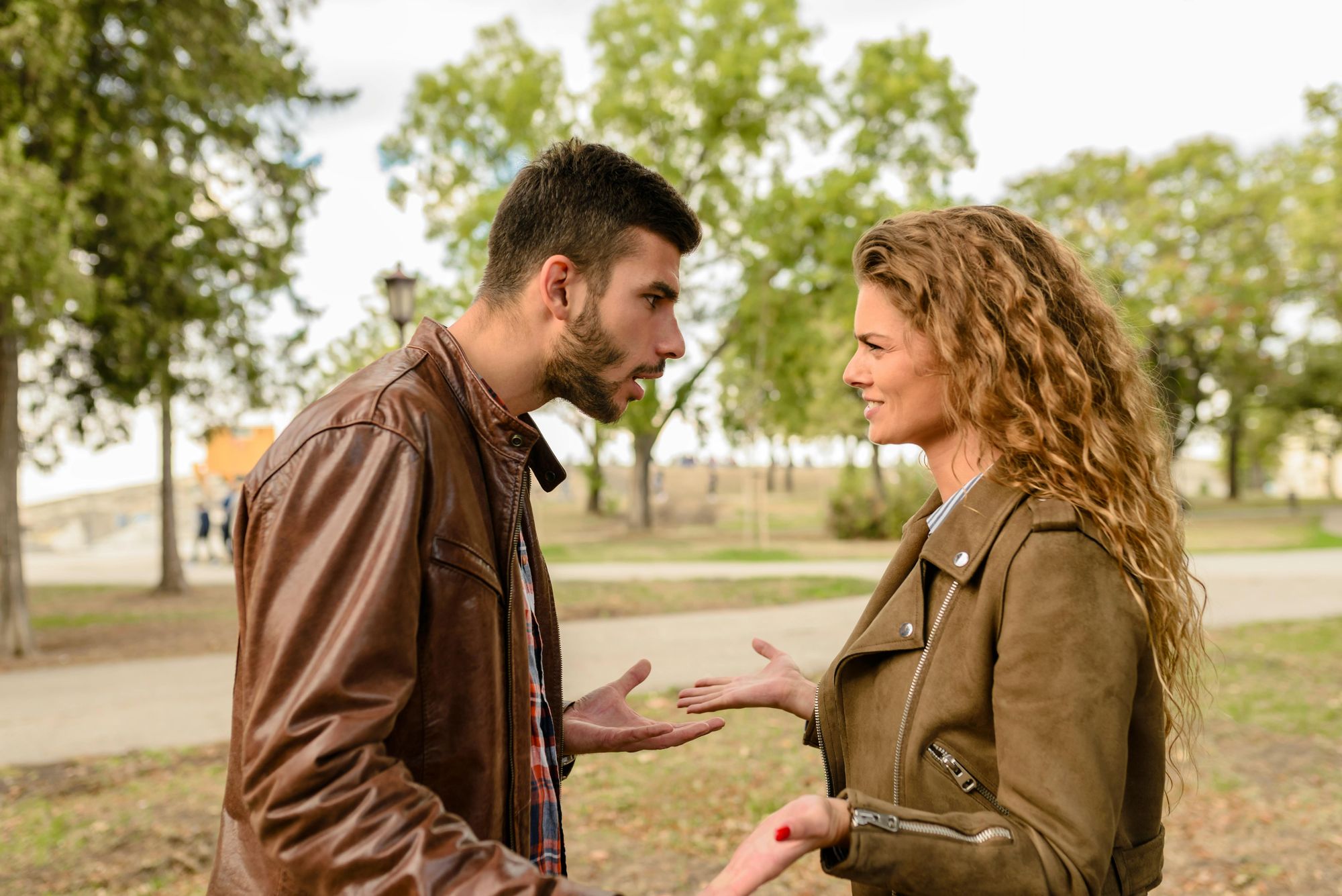 Two caucasian persons facing each other and arguing. One has short dark hair and facial hair with plaid button down and leather jacket. Other has long curly hair with striped button down and brown suede jacket.