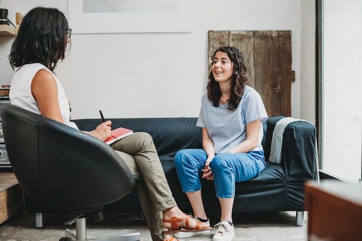 one adult on a couch speaking happily with professional adult sitting in a chair across from them