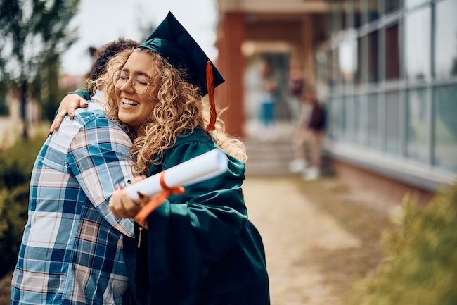 a curly-haired teenager wearing a cap and gown hugging a loved one