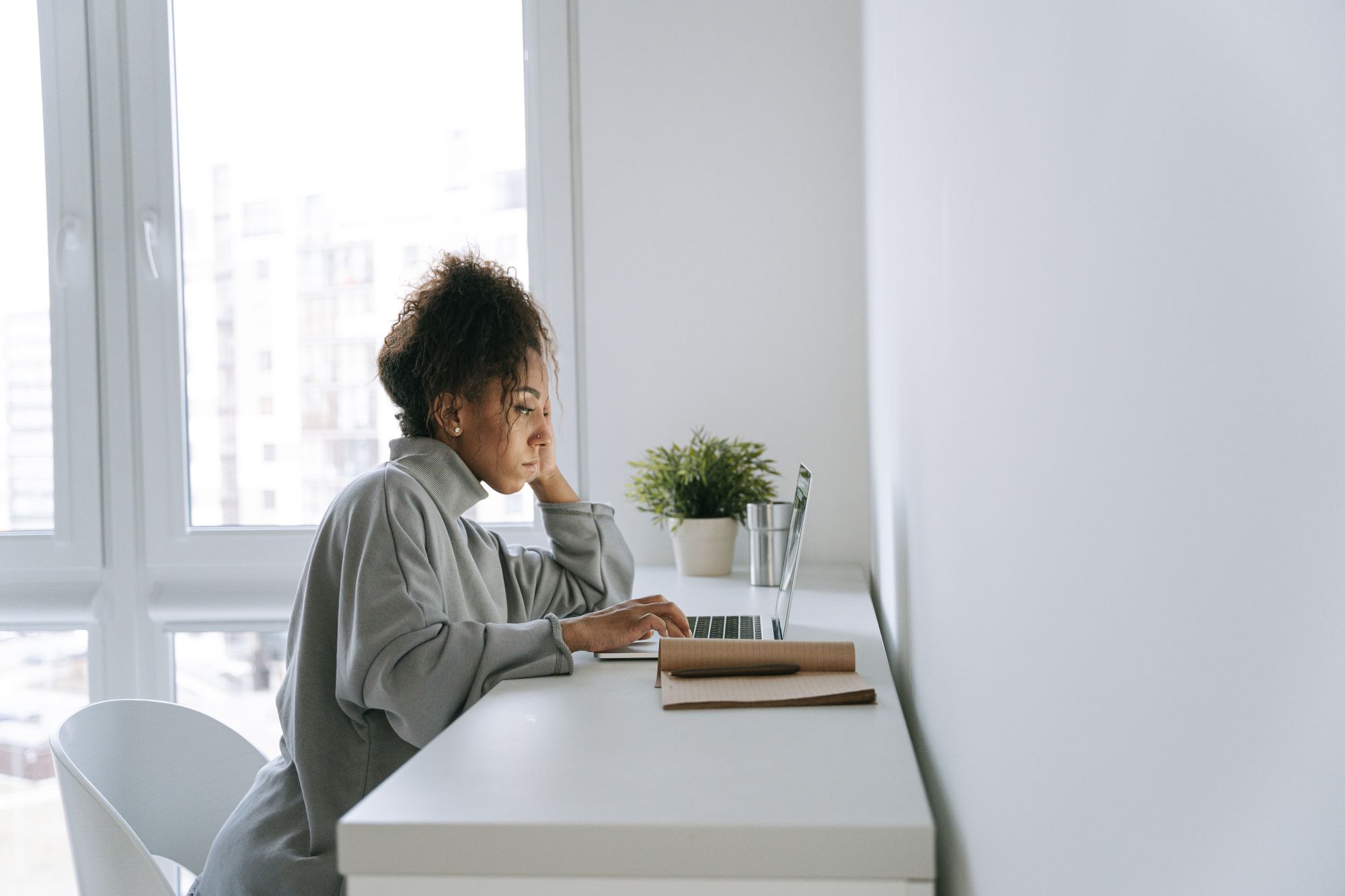 Dark skinned woman sitting at a white desk, working at a laptop.